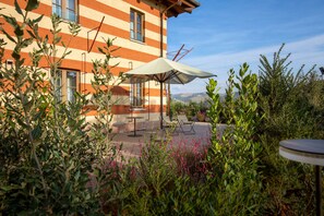 A building with striped exterior walls, a patio area with outdoor furniture, and surrounding greenery. - Villaggio Narrante - Cascina Galarej (Serralunga d'Alba)