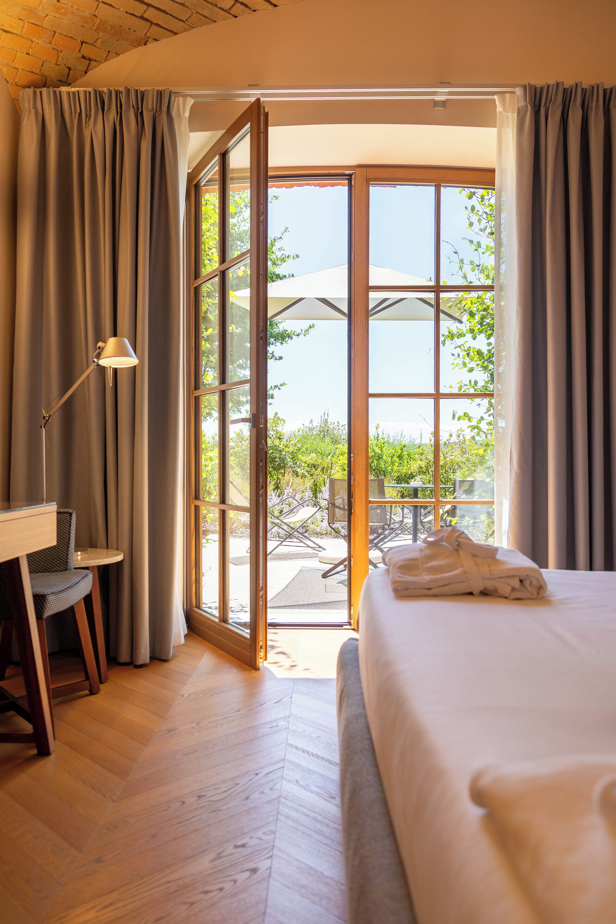Habitación de hotel con cama, escritorio, silla y vista a través de un ventanal a un espacio exterior con mesa y sillas.