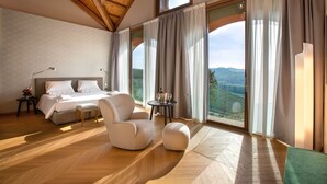 Un dormitorio con una cama grande, una zona de estar con una silla y reposapiés, una mesita de noche con lámpara y vistas al campo a través de amplios ventanales.