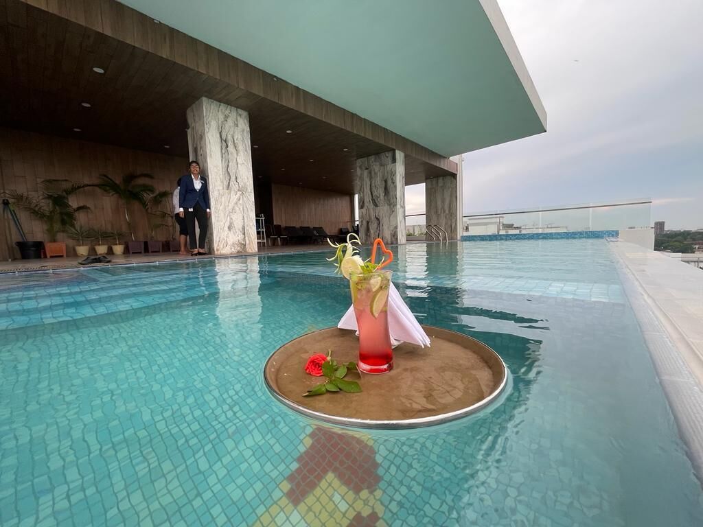 Zona de piscina con una bebida en una bandeja, una rosa y una persona de fondo.