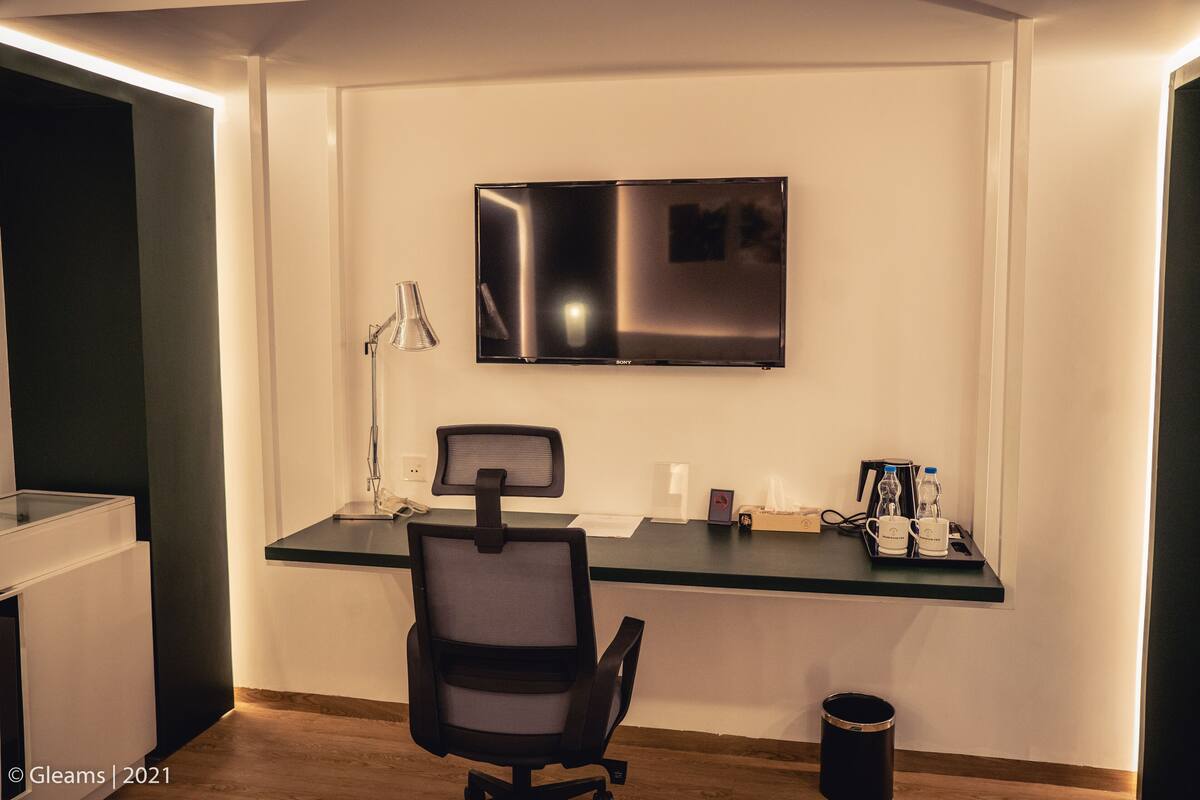A modern hotel room with a flat-screen TV mounted on the wall, a desk with a chair, and a small trash bin.