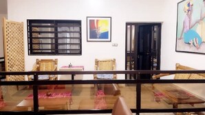 Una habitación con una mesa de comedor, dos sillas de mimbre, una ventana con persianas, un cuadro abstracto enmarcado y una puerta.