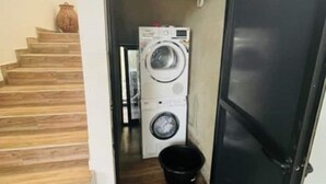Un lavadero con lavadora y secadora, un cubo negro y una escalera de madera.