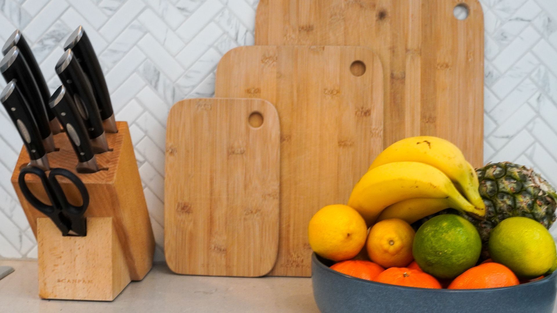 Una encimera de cocina con un frutero que contiene plátanos, limones, limas y naranjas, un par de tijeras y un bloque de cuchillos con varios cuchillos.