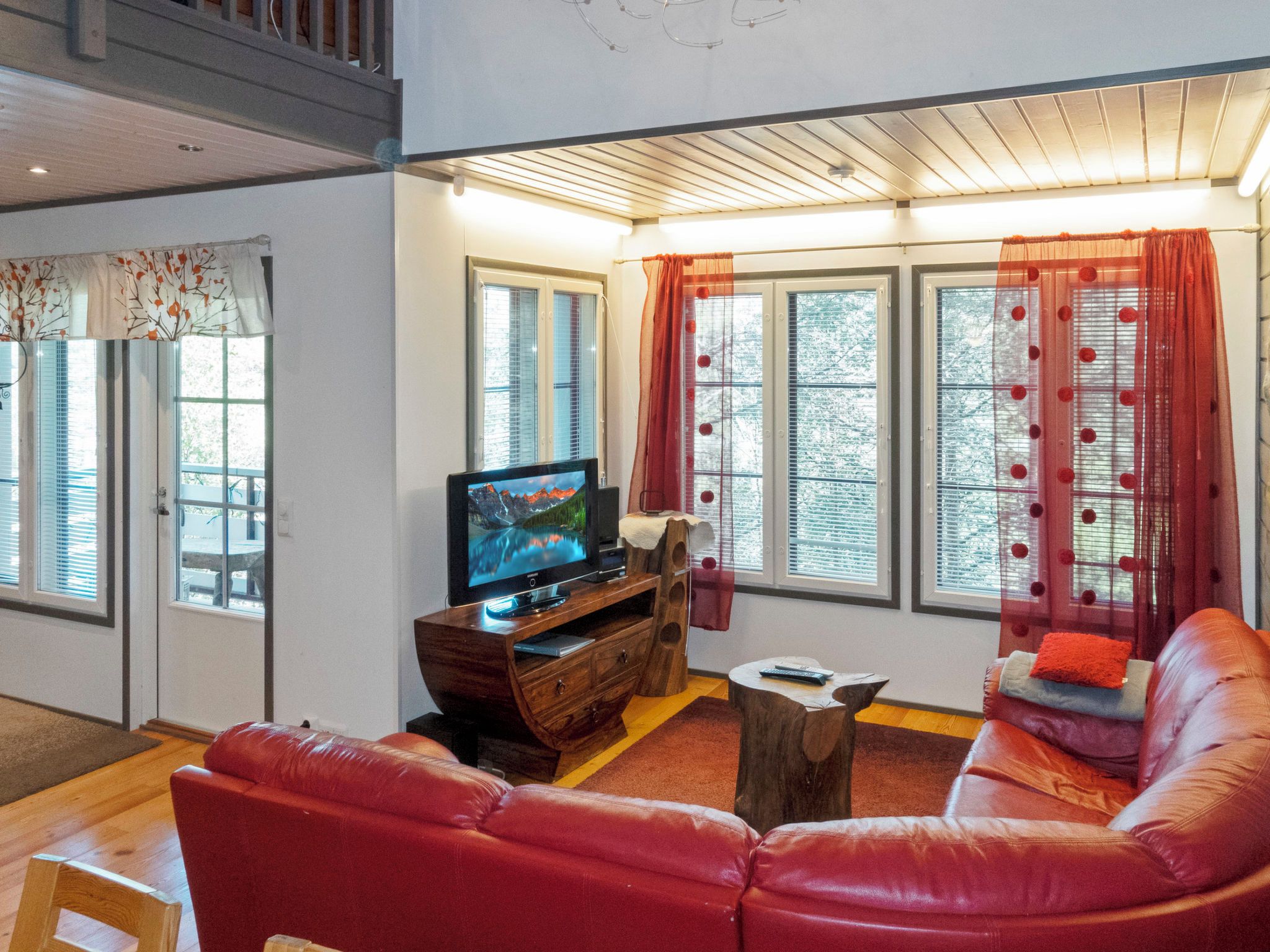 A living room with a red leather sofa, a wooden entertainment unit with a TV, and large windows with curtains.