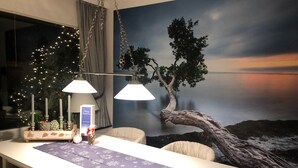 A dining area with a table set for two, a mural of a tree and sunset, and a Christmas tree with lights. - Vacation home with 80 sqm - on Ballumweg on Sylt (Sylt)