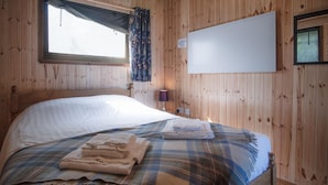 Quarto com painéis de madeira, cama, janela com cortinas, televisão na parede e uma luminária.