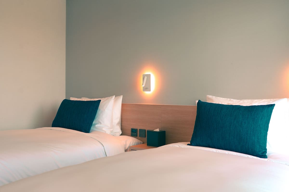 A hotel room with two beds, a nightstand, a wall-mounted light, and a wooden headboard.
