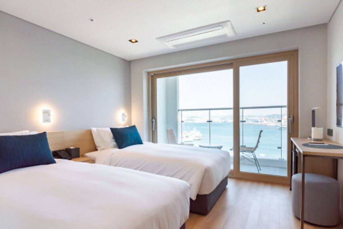 A modern hotel room with two beds, a desk, and a large sliding glass door leading to a balcony with a city view.