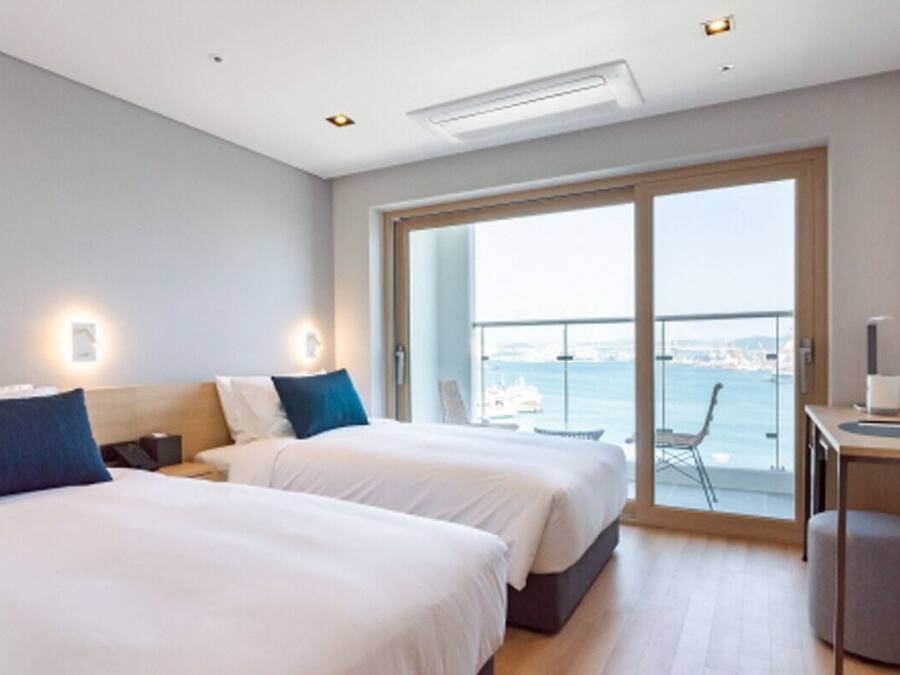 A modern hotel room with two beds, a desk, and a large sliding glass door leading to a balcony with a city view.