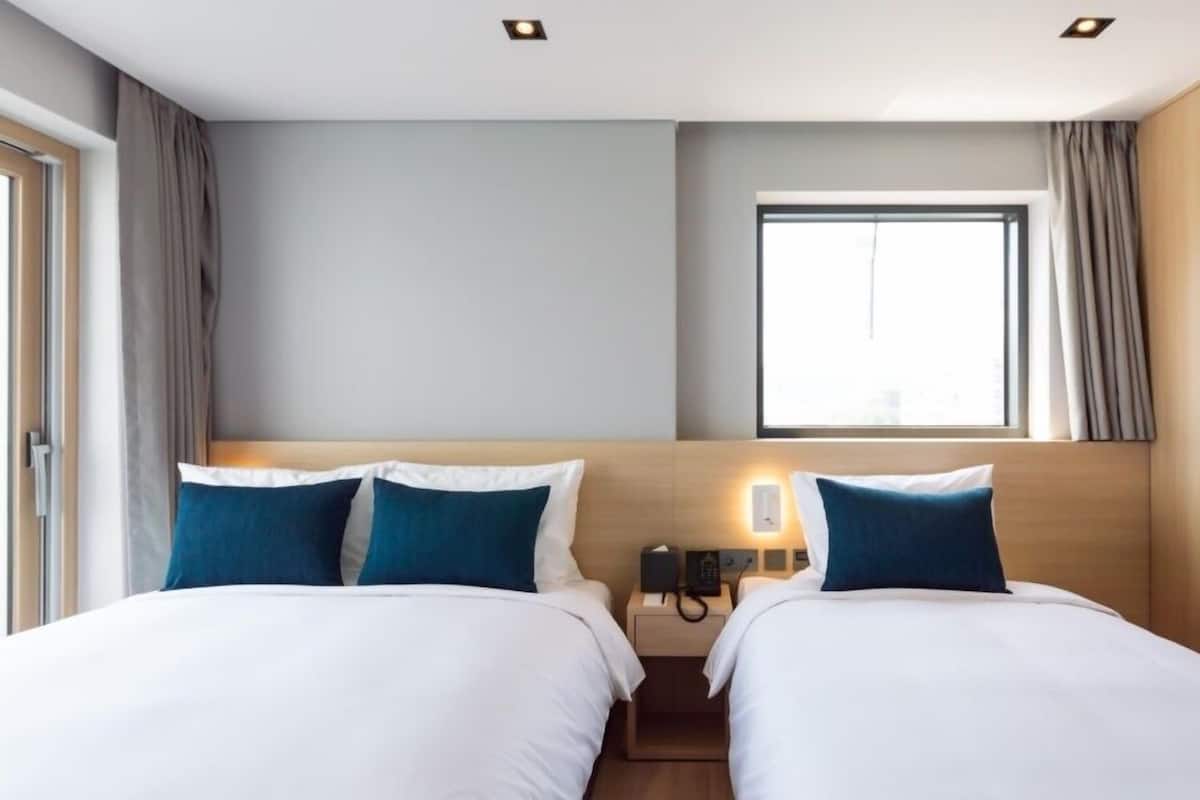 A hotel room with two beds, a bedside table with a phone, a window with curtains, and a light fixture on the ceiling.