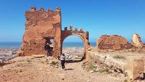 Une structure en pierre d’époque, dotée d’une arche, entourée d’un paysage rocheux et offrant une vue lointaine sur une ville côtière.