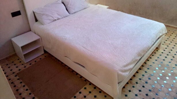 Un lit simple avec une tête de lit et un pied de lit blancs, une petite table de chevet et un tapis à motifs posé sur un sol carrelé.