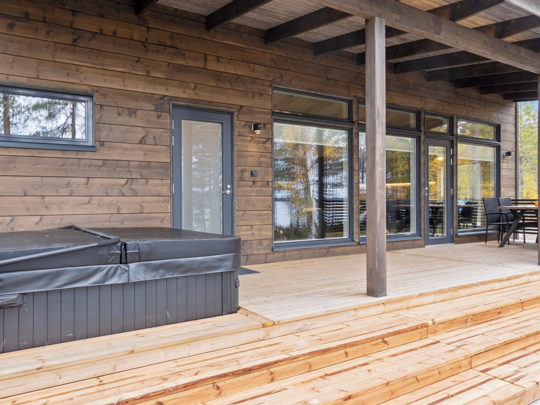 Eine Holzterrasse mit Whirlpool, großen Fenstern und einer überdachten Sitzgelegenheit.