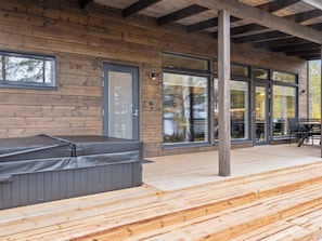 Eine Holzterrasse mit Whirlpool, großen Fenstern und einer überdachten Sitzgelegenheit.