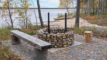 Leirintäalue järven rannalla, jossa on kivistä rakennettu nuotiopaikka, puinen penkki ja kanootti.