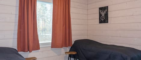 Huoneessa on kaksi sänkyä, ikkuna, jossa on verhot, pieni jakkara ja seinälle ripustettu taulu.