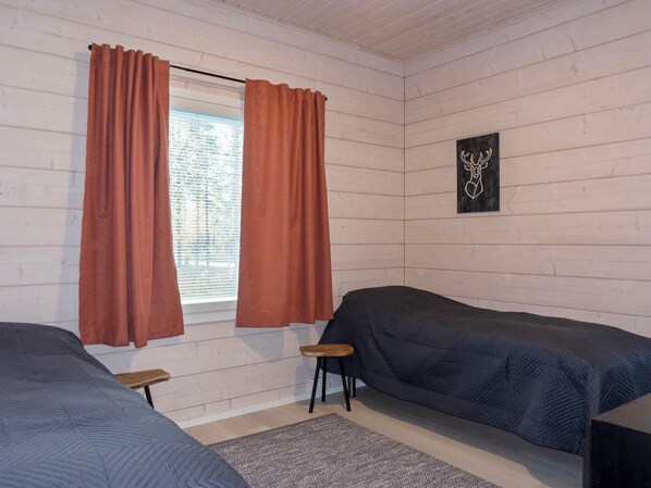 Huoneessa on kaksi sänkyä, ikkuna, jossa on verhot, pieni jakkara ja seinälle ripustettu taulu.