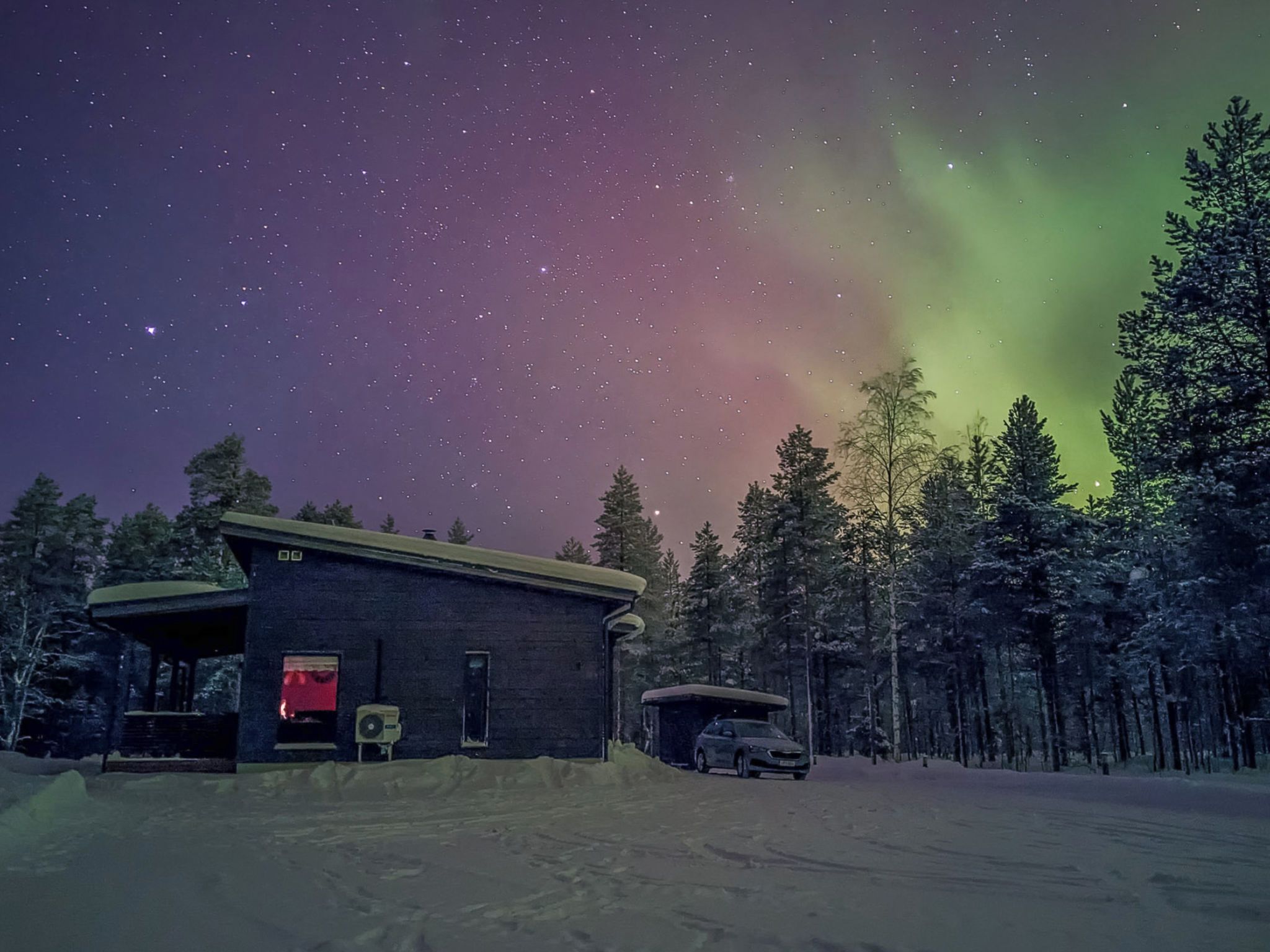 Une cabane entourée de neige, une voiture et une forêt en arrière-plan.