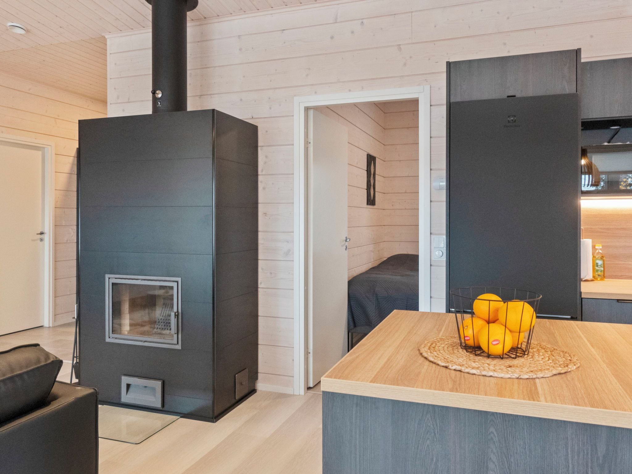 Un interior moderno con un estufa de madera negra grande, una mesa de comedor de madera con naranjas, y una zona de cocina.