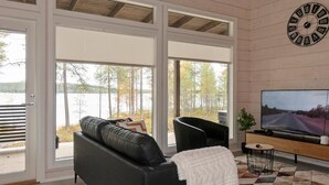 Ein modernes Wohnzimmer mit einem schwarzen Ledersofa, einem Fernseher und einem großen Fenster mit Blick auf Bäume und einen See.