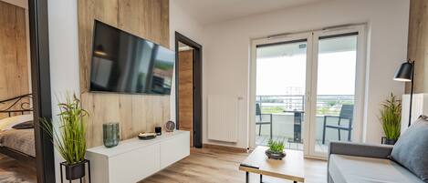 A modern living room with a flat-screen TV, wooden panel walls, a sofa, a dining table, and a balcony with a view.
