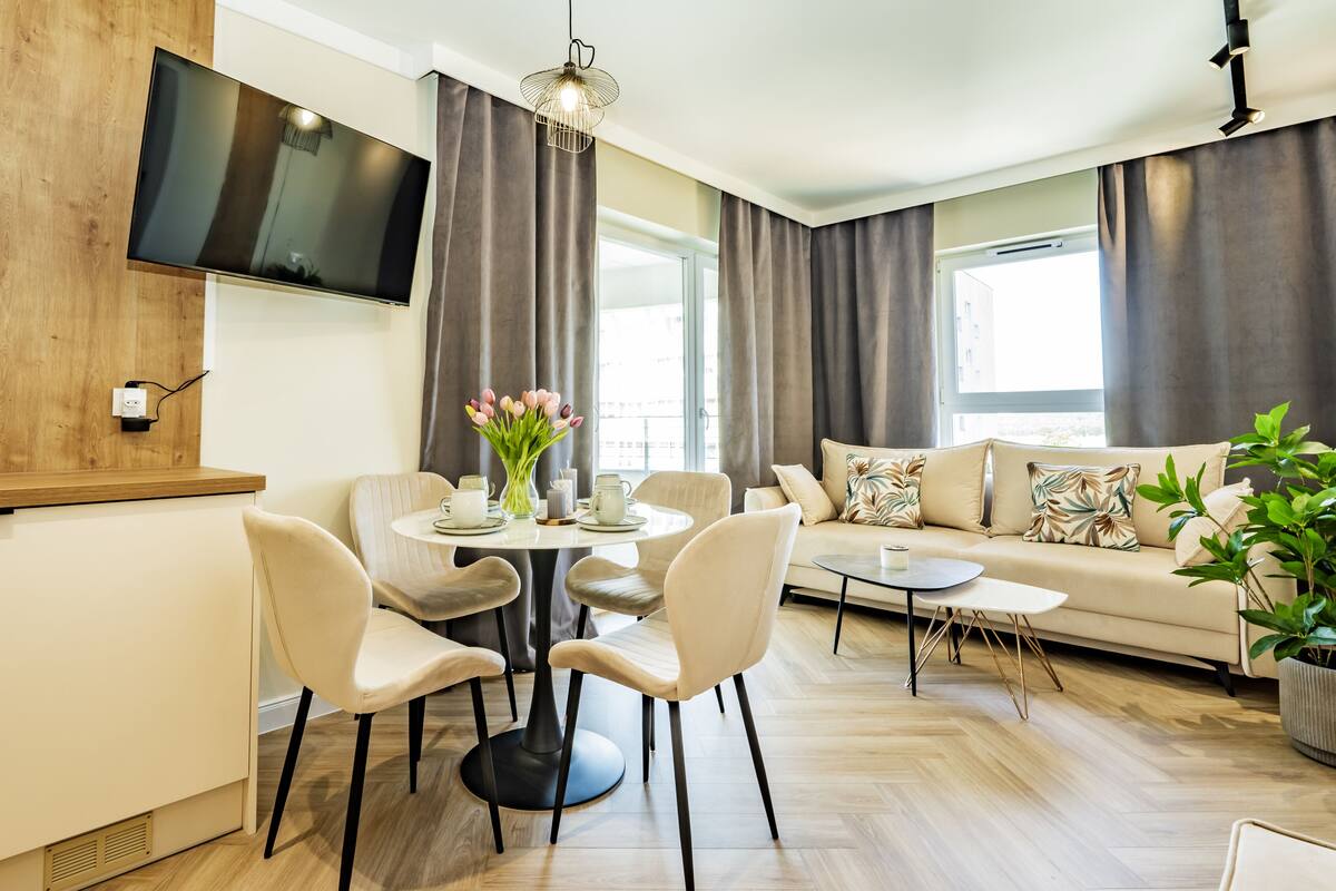 Een moderne woonkamer met een bank, een eettafel met stoelen, een aan de muur bevestigde televisie en een groot raam met gordijnen.