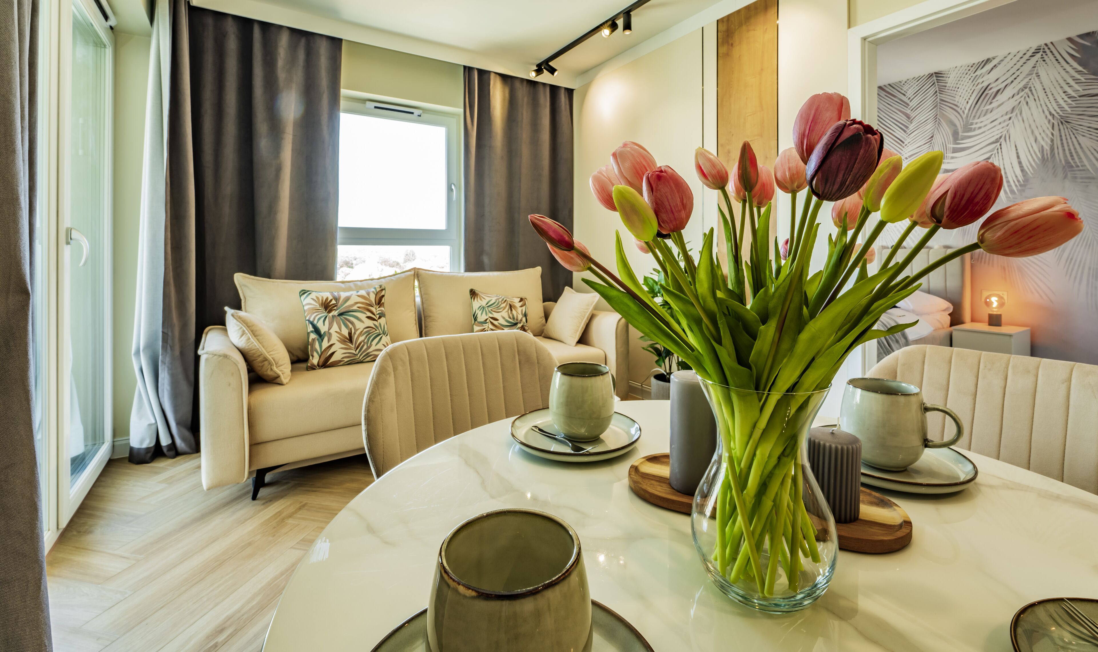 A modern living room with a beige sofa, a glass vase with green plants, and a dining table with plates and cups.