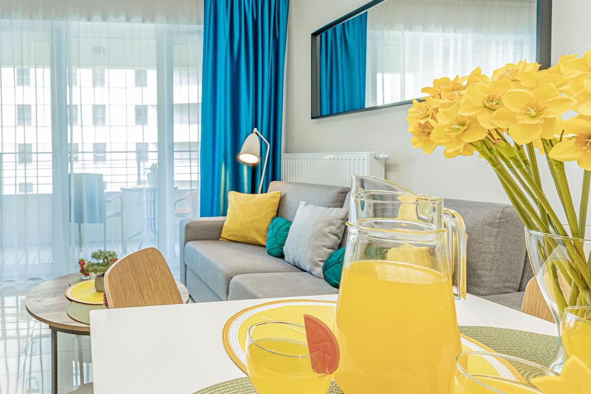Een moderne woonkamer met een bank, een eettafel met gele placemats en een groot raam met blauwe gordijnen.