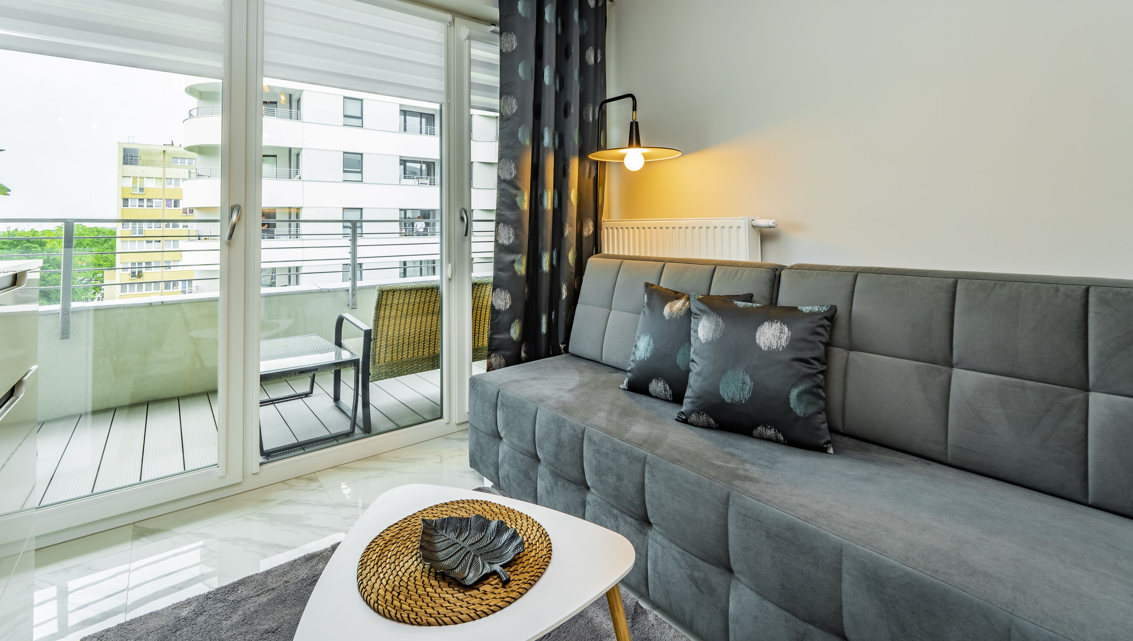 En moderne stue med en grå sofa, et lille hvidt bord og en flettræbakke med bladmotiv. En sort gardin med bladmotiv er delvist trukket til side, og der står en gulvlampe tændt.