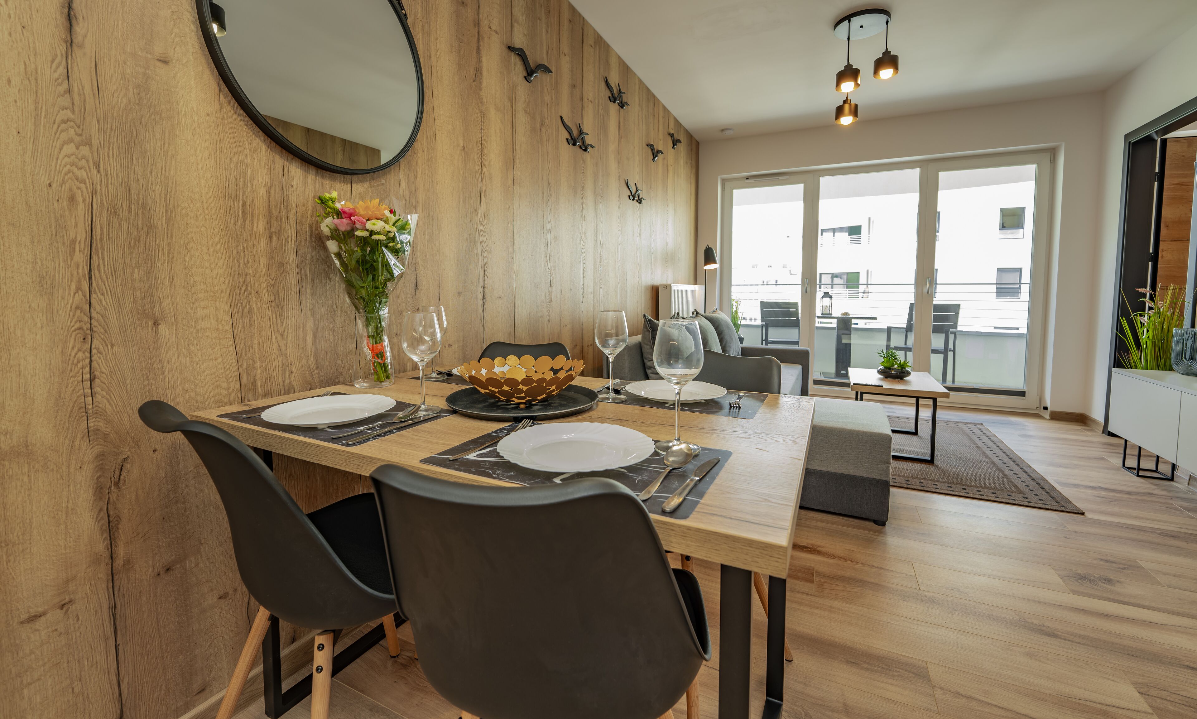 A dining area with a wooden table set for four, chairs, a vase with flowers, and a view of the city through large windows.