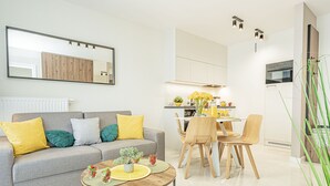 Een moderne woonkamer met een grijze bank, gele en groene kussens, een salontafel met decoratieve objecten, en een eethoek met houten stoelen en een witte tafel.