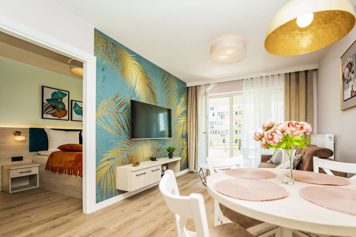 Een moderne woonkamer met een grote flatscreen-tv, een eethoek met een ronde tafel en roze placemats, en een levendig, tropisch blad-muurschildering.