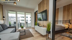 Een modern appartement met een grijze bank, een kleine tafel met een plant, een wit kastje en een bed met een grijze deken.