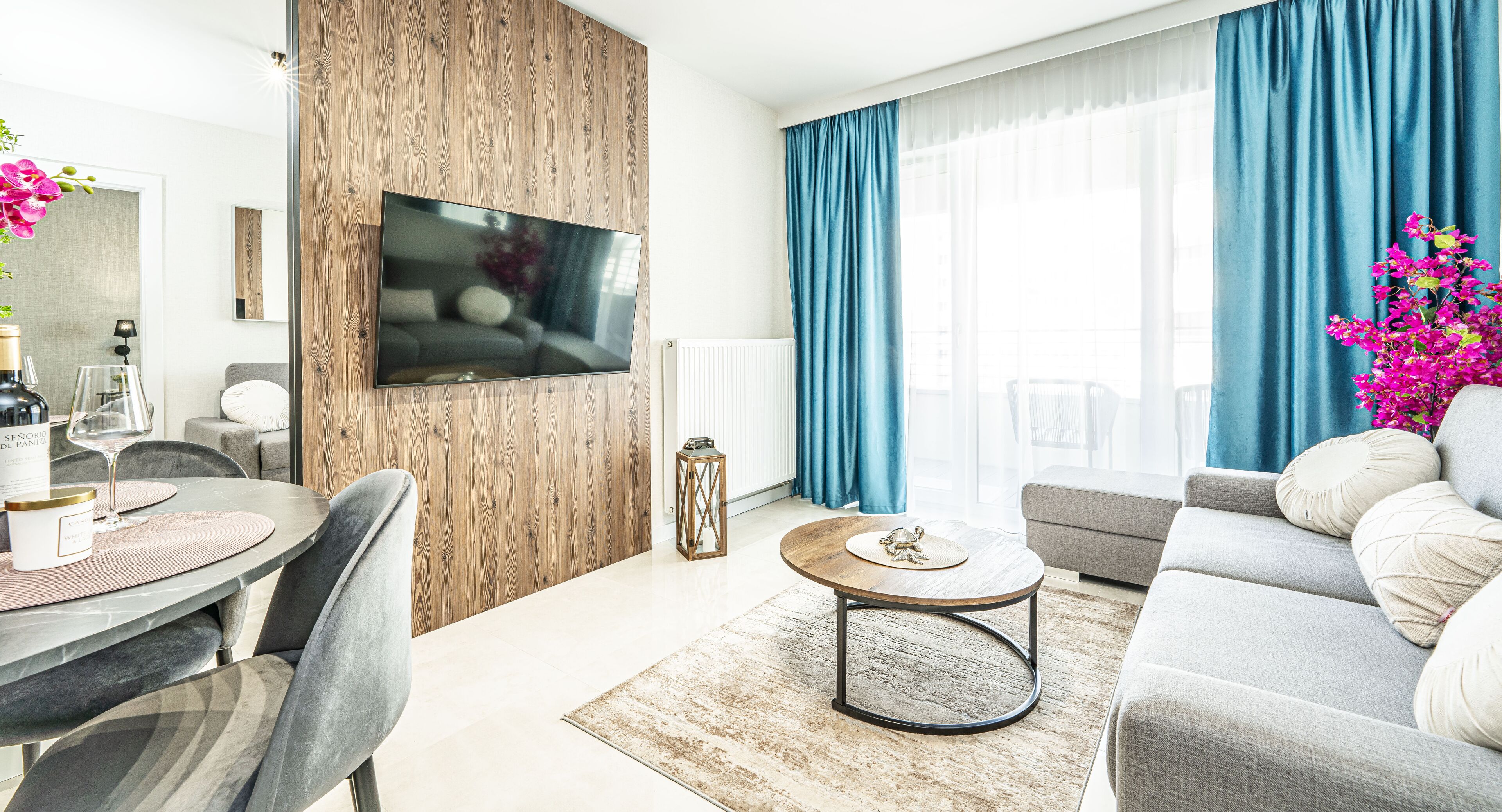 A modern living room with a flat-screen TV mounted on a wooden wall, a grey sofa, a round coffee table, and a dining area with a table and chairs.