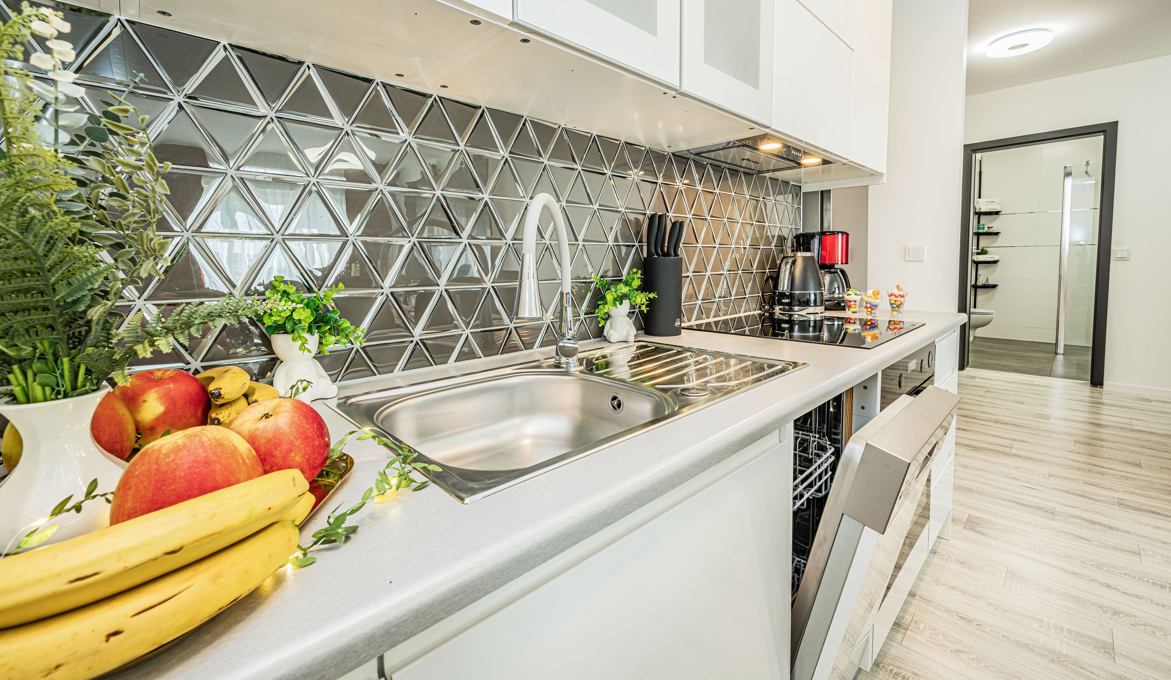 A modern kitchen with a stainless steel sink, a geometric backsplash, and a variety of fruits and plants on the counter.