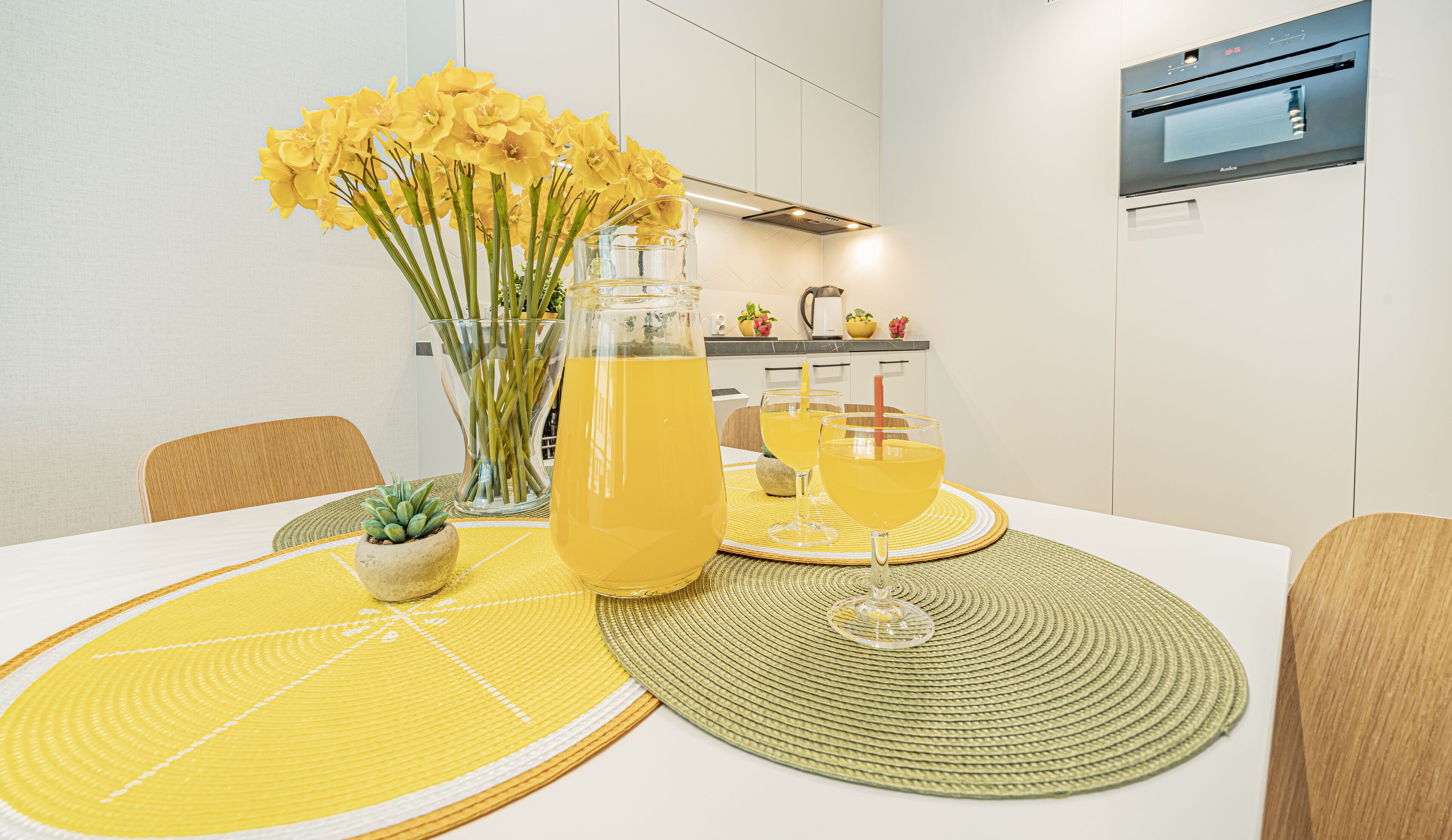 Une table à manger avec des sets de table jaunes, un vase rempli de fleurs jaunes et une carafe contenant une boisson jaune.