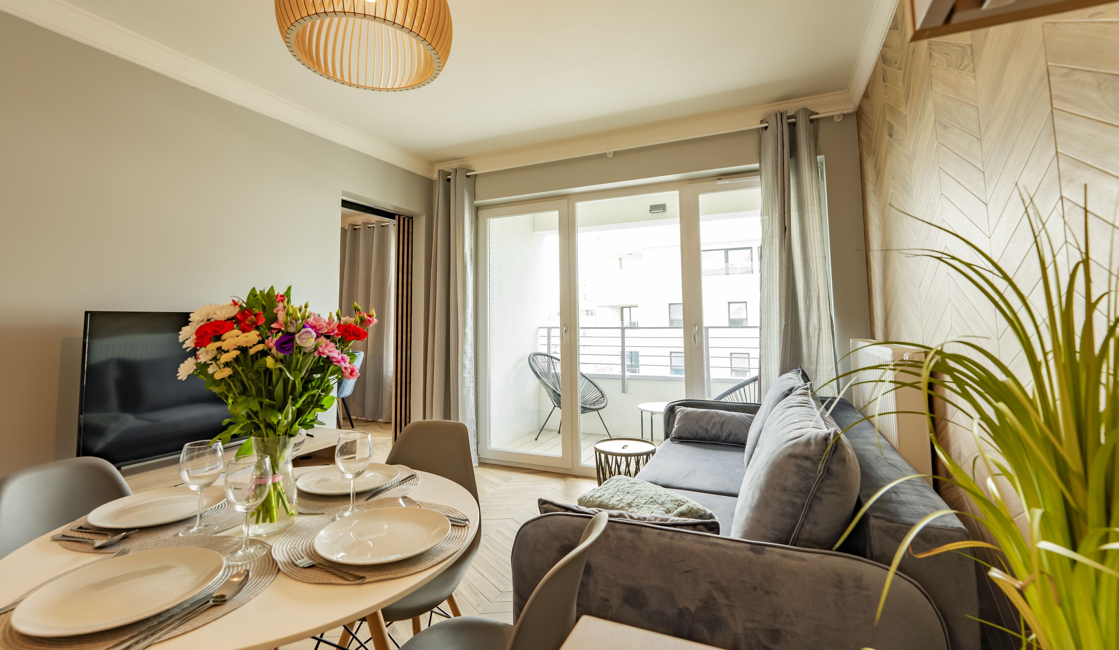 A modern living room with a dining area, a sofa, a TV, and a balcony with a view of buildings.