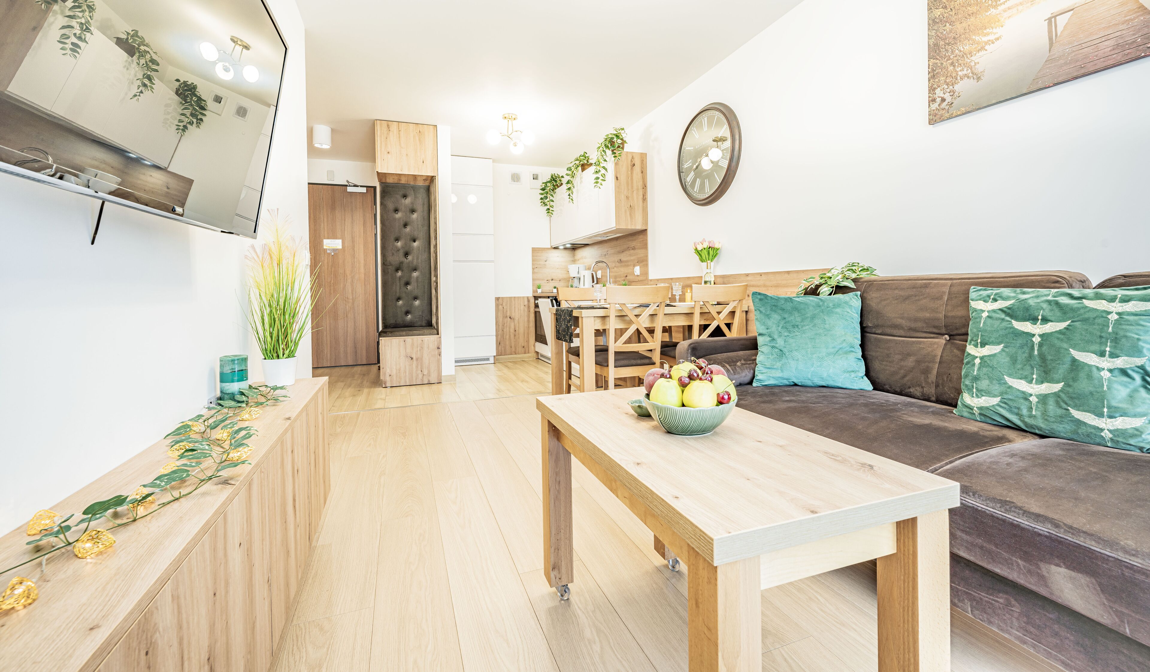 A modern living room with a wooden coffee table, a brown sofa with a teal cushion, and a kitchen in the background.