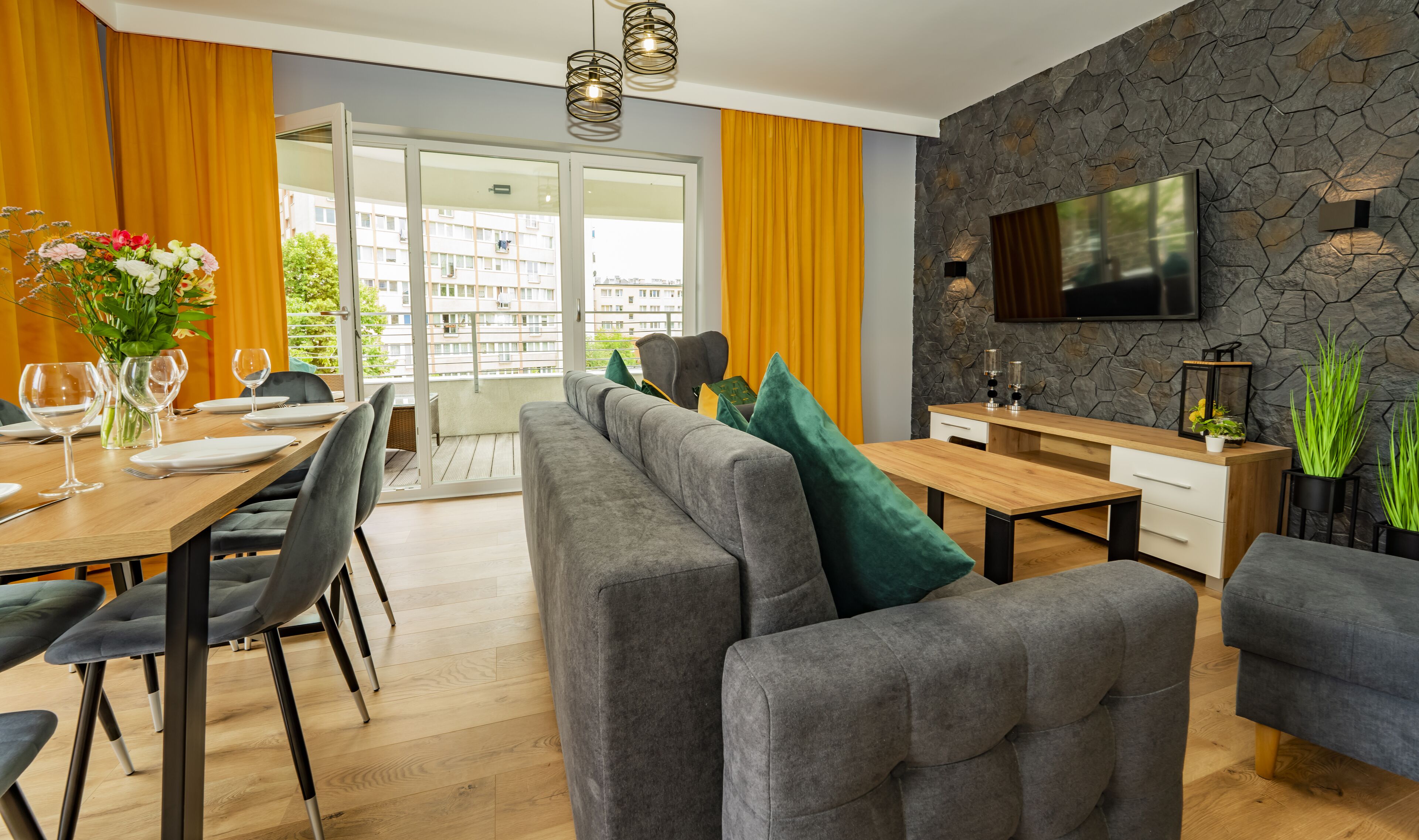 A modern living room with a dining area, grey sofas, a wooden dining table, and a stone accent wall with a TV.