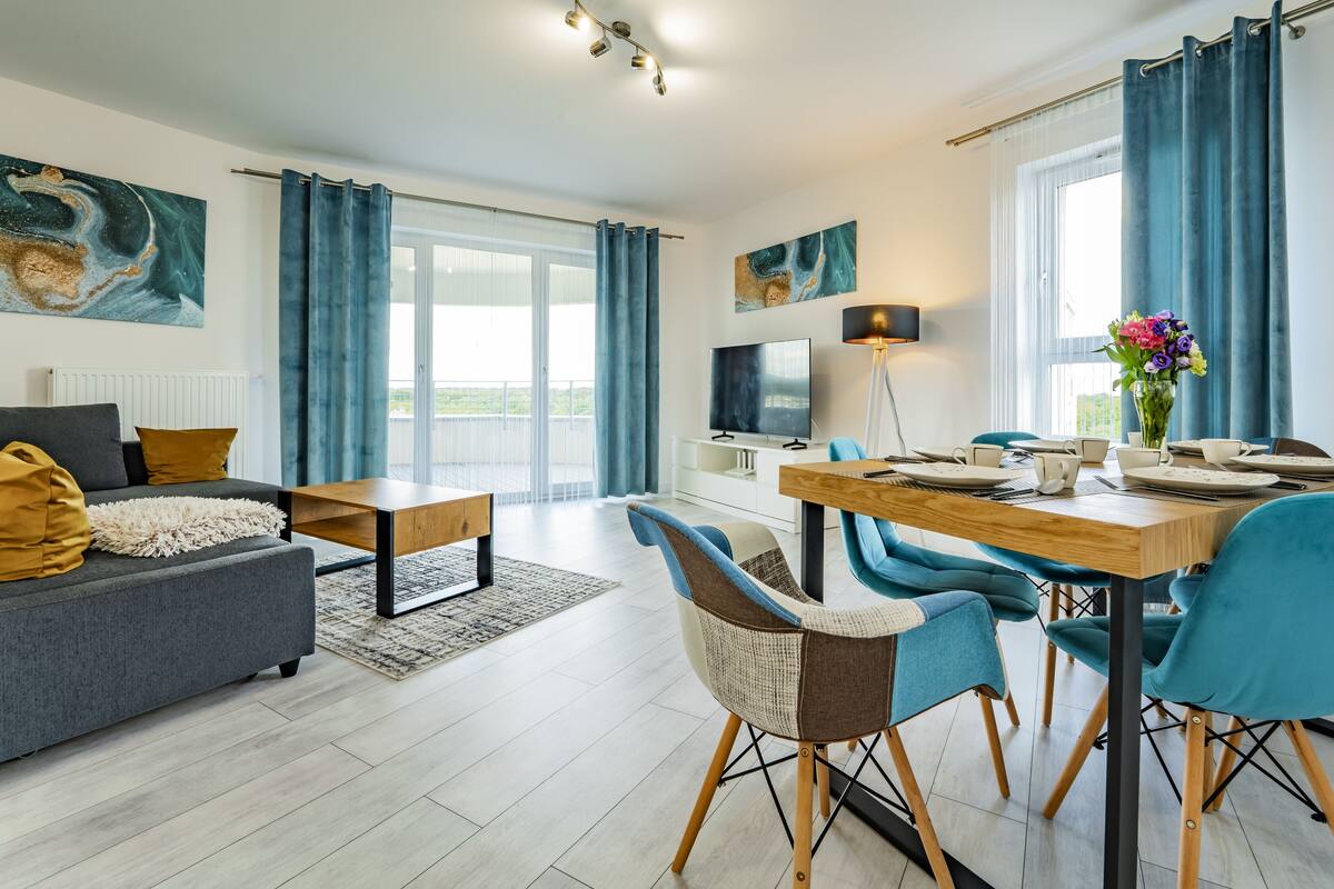 Een moderne woonkamer met een grijze bank, een houten salontafel en een eethoek met blauwe stoelen en een houten tafel.