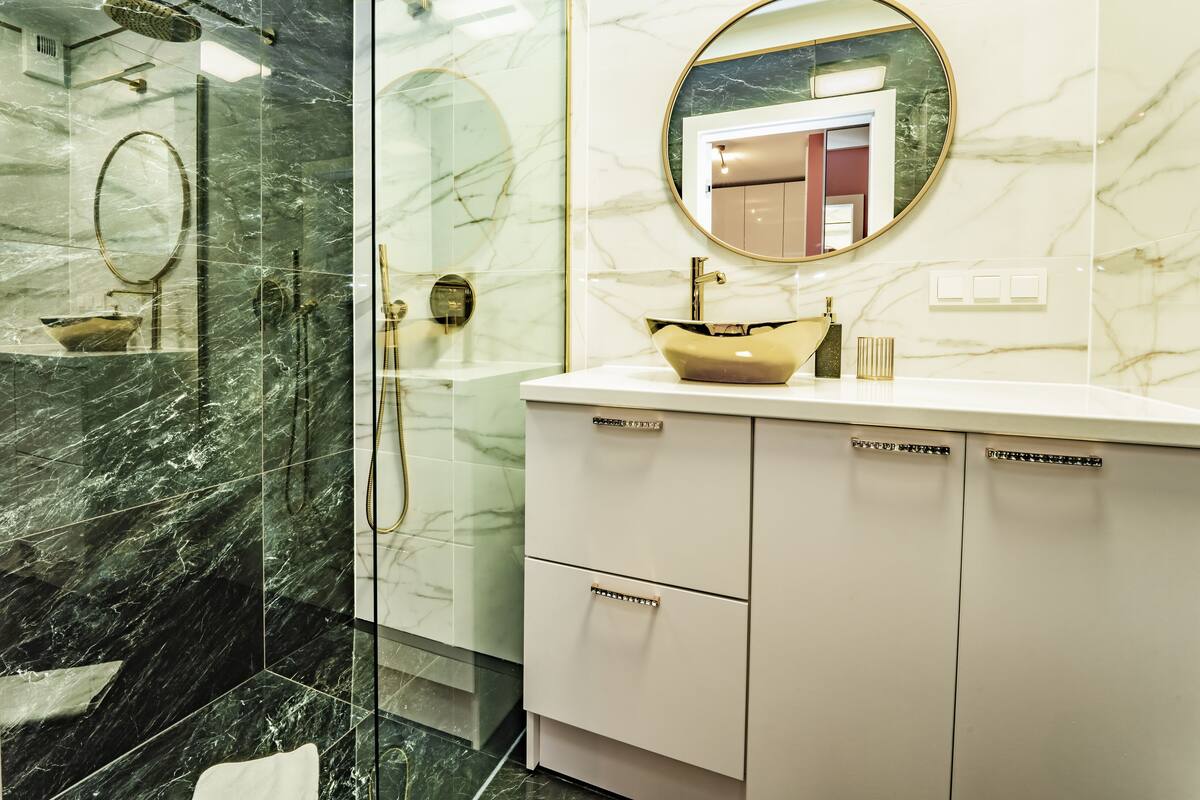 Een badkamer met een marmeren vloer, een glazen douchecabine, een wit wastafelmeubel met een gouden wastafel en een spiegel met een gouden lijst.