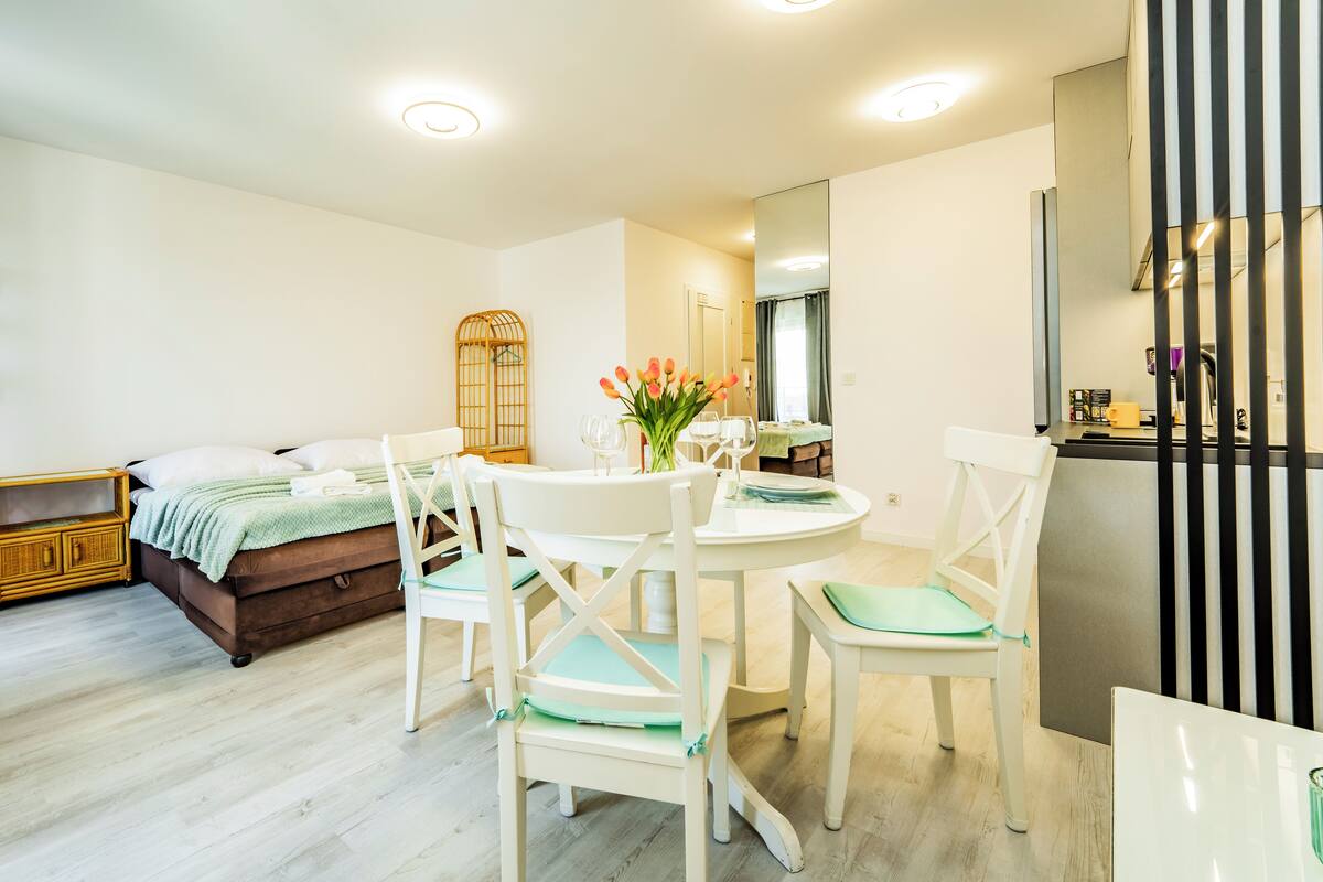 Een moderne, goed verlichte kamer met een bed, een eettafel met stoelen, een klein keukentje en een boekenkast.