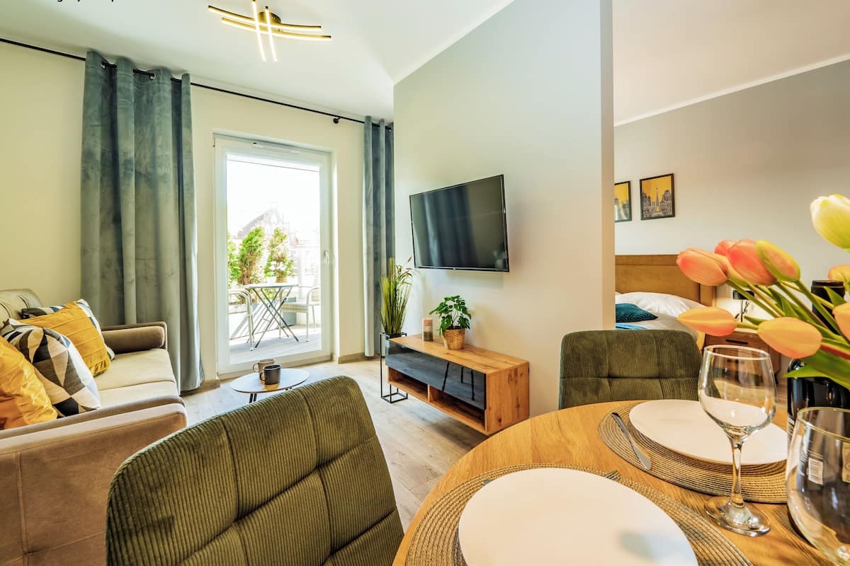 Een moderne woonkamer met een bank, fauteuils, een eettafel gedekt voor twee personen, een flatscreen televisie en een balkon met uitzicht op bomen en gebouwen.