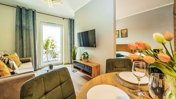 A modern living room with a sofa, armchairs, a dining table set for two, a flat-screen TV, and a balcony with a view of trees and buildings.