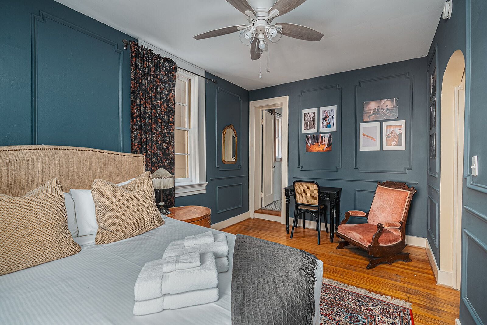 A bedroom with a bed, a desk, two chairs, a ceiling fan, and a door leading to another room.