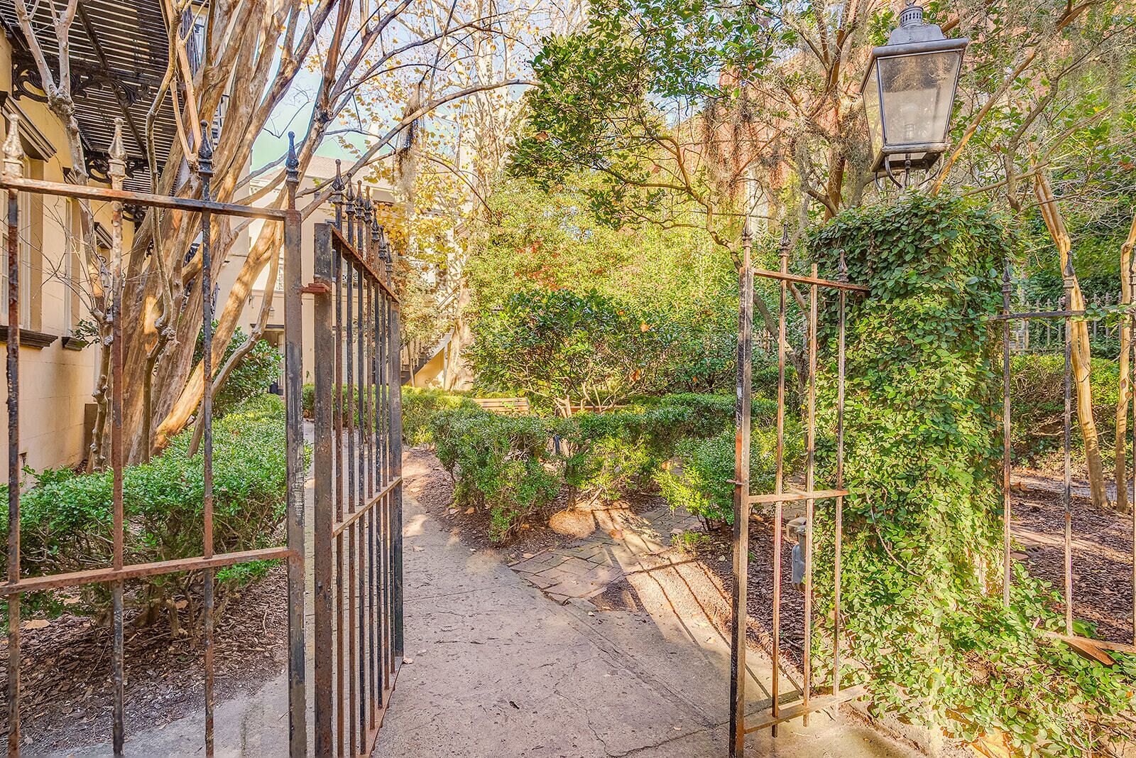 A narrow pathway flanked by metal fences and lush greenery, leading to a building with a traditional architectural style.