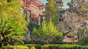A brick building with arched windows, surrounded by lush greenery and trees.