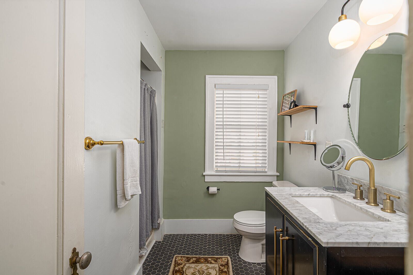 A bathroom with a toilet, sink, and mirror. There is a window with blinds, a towel rack, and a rug on the floor.