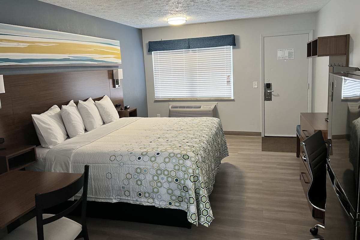 Quarto de hotel com cama, escrivaninha, cadeira e uma grande janela com persianas.